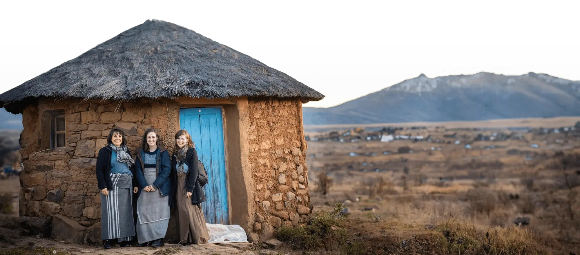 Three women in front of a house on a frontier with mountains in the background